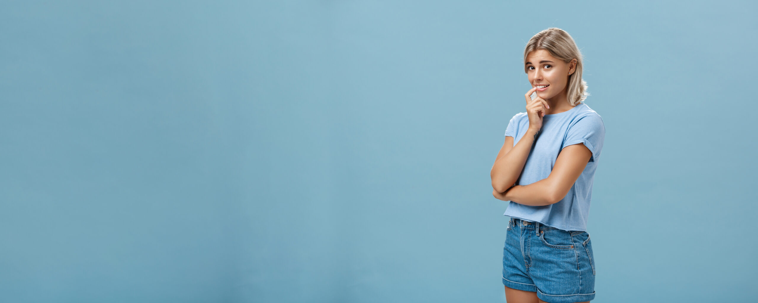 studio shot unconfident silly insecure cute feminine girl with blond hair standing halfturned blue background holding finger lip while being shy ask question smiling hesitating scaled